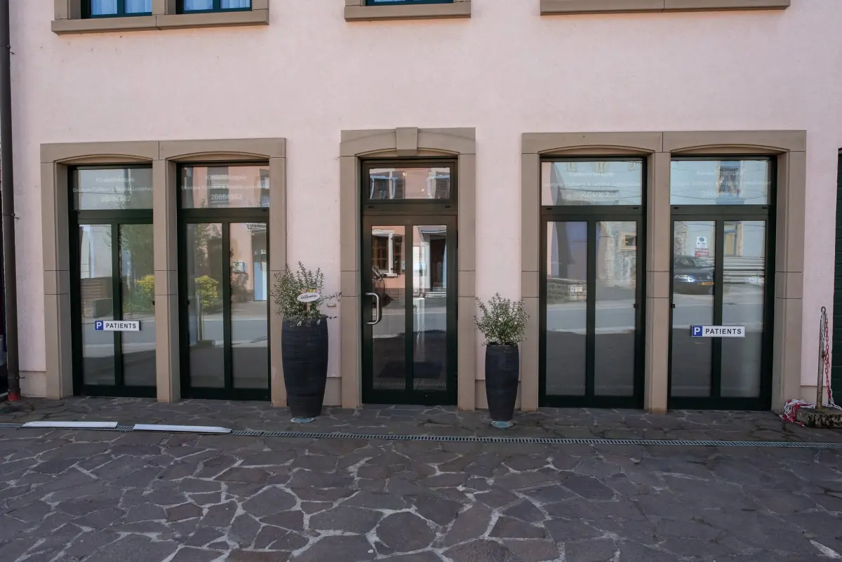 Der Eingang der Praxis: rosa Fassade, dunkle Glastüren, Schilder „P Patients“ und zwei Pflanzkübel