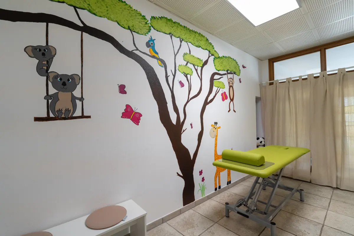 Der Kinderraum: ein gemalter Baum mit Koalas auf einer Schaukel, Giraffe und Papagei, daneben eine grüne Behandlungsliege